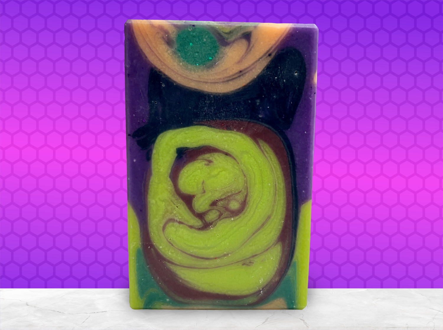 A rectangular handmade soap bar with a marbled swirl pattern of lime green, purple, black, and orange, standing upright on a white marble surface against a purple and pink patterned background.