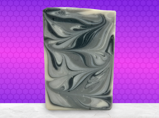 A rectangular bar of soap with a marbled swirl pattern of black, gray, and off-white, standing upright on a white surface against a purple and pink honeycomb-patterned background.