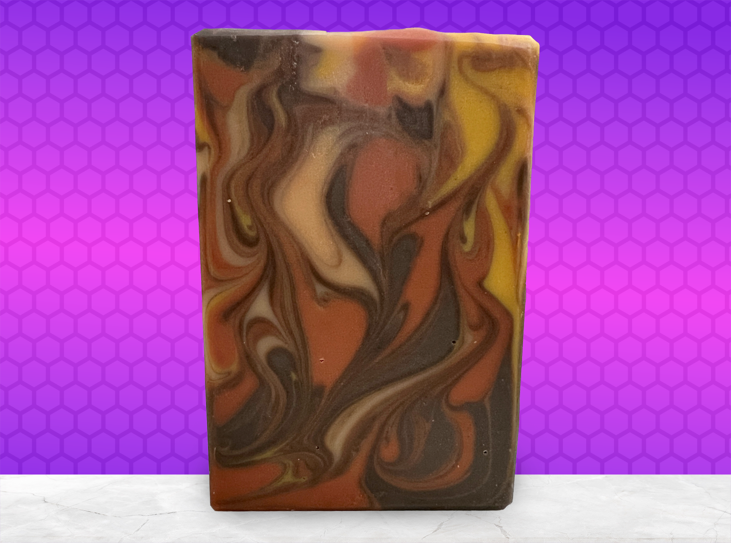 A rectangular bar of marbled artisan soap with swirls of orange, brown, cream, and yellow, standing upright on a white marble surface against a purple and magenta background.