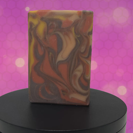 A rectangular bar of marbled artisan soap with swirls of orange, brown, cream, and yellow, standing upright against an animated purple and magenta background.