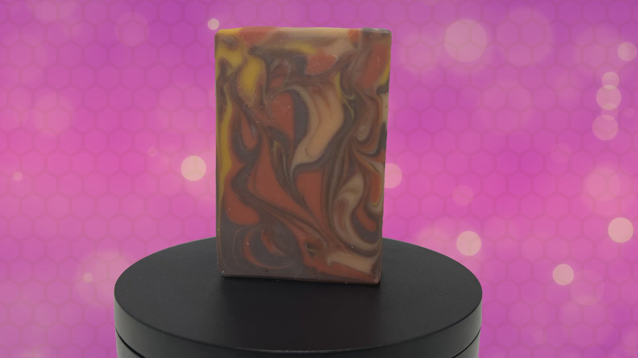 A rectangular bar of marbled artisan soap with swirls of orange, brown, cream, and yellow, standing upright against an animated purple and magenta background.