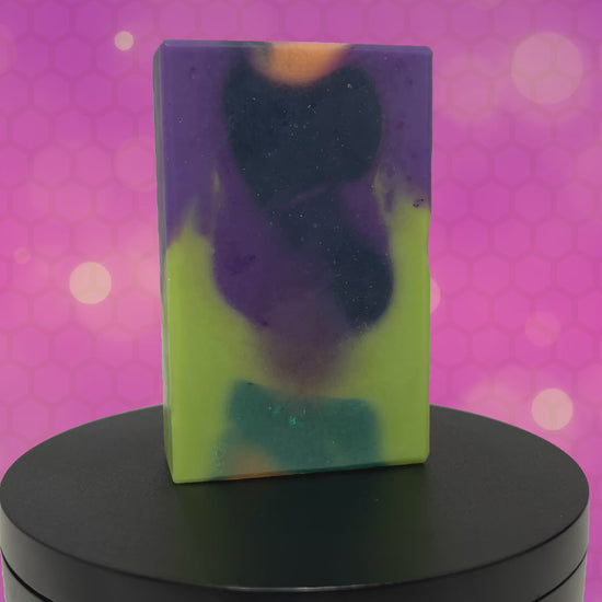 A rectangular handmade soap bar with a marbled swirl pattern of lime green, purple, black, and orange, standing upright  against an animated purple and pink patterned background.