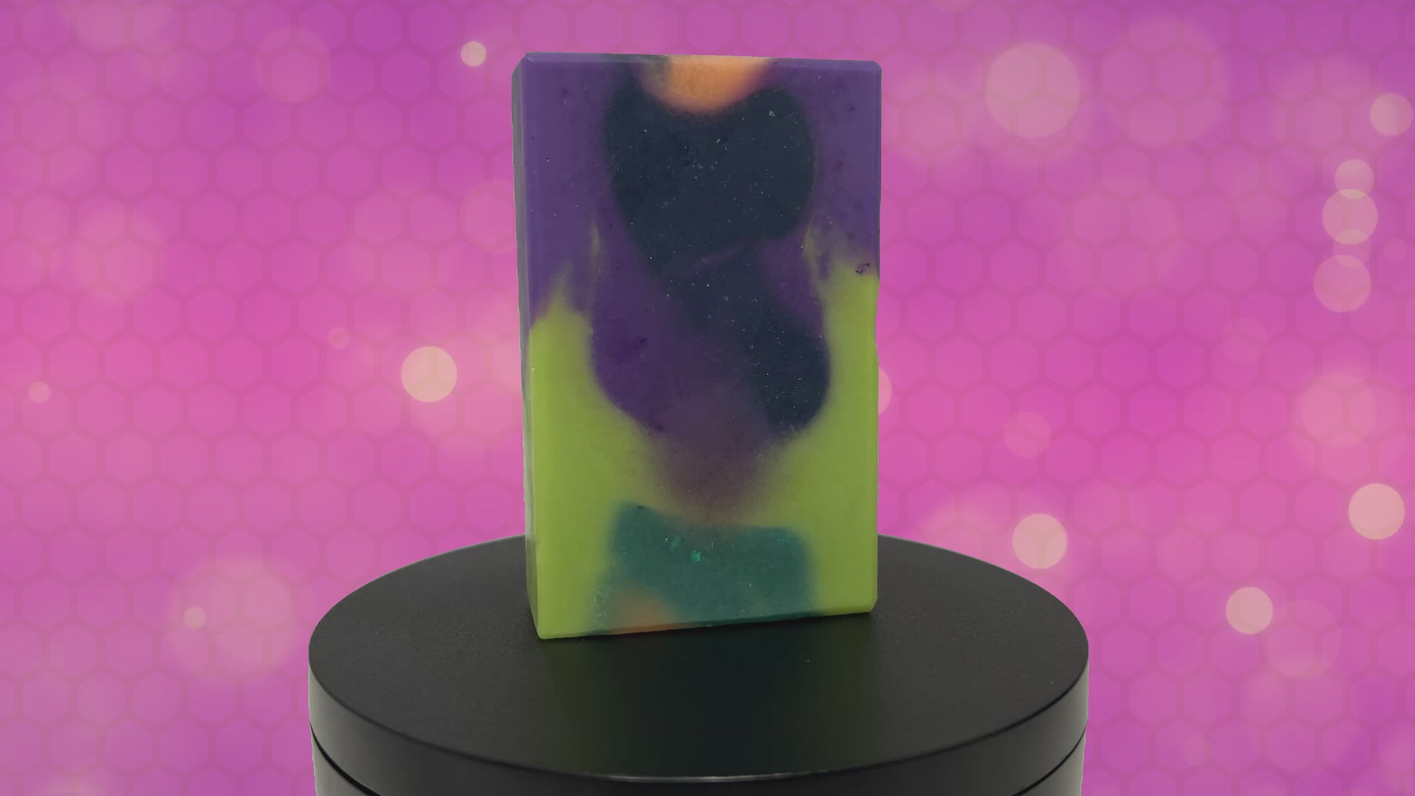 A rectangular handmade soap bar with a marbled swirl pattern of lime green, purple, black, and orange, standing upright  against an animated purple and pink patterned background.