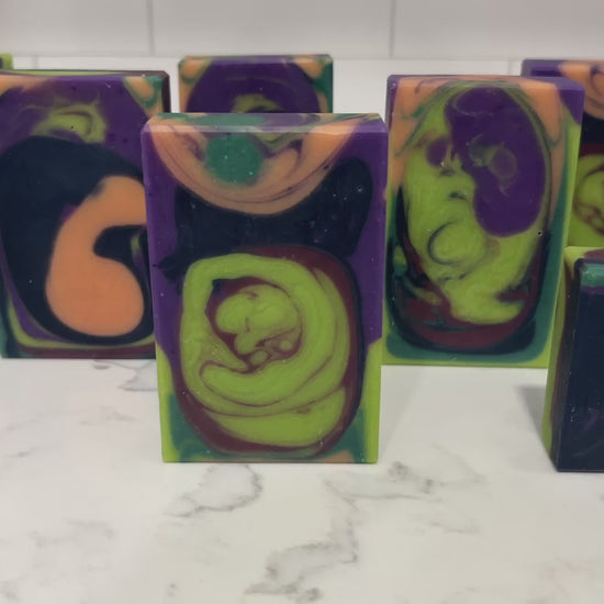 Rectangular handmade soap bars with a marbled swirl pattern of lime green, purple, black, and orange, standing upright on a white marble surface against a purple and pink patterned background.