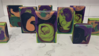 Rectangular handmade soap bars with a marbled swirl pattern of lime green, purple, black, and orange, standing upright on a white marble surface against a purple and pink patterned background.