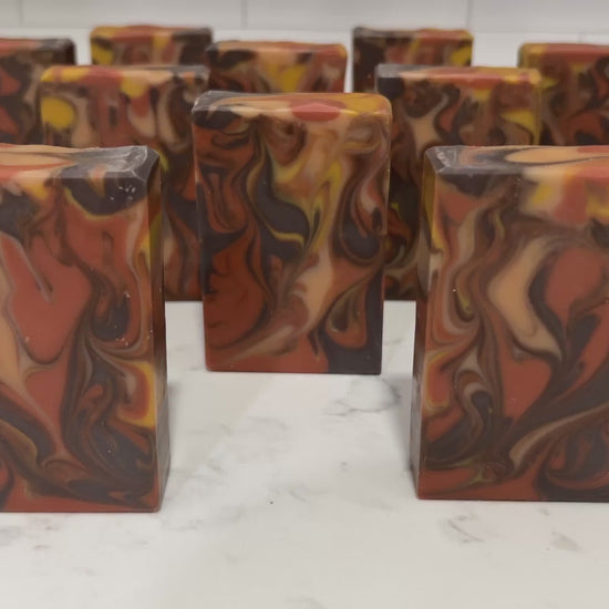 Rectangular bars of marbled artisan soap with swirls of orange, brown, cream, and yellow, standing upright on a white marble surface against a purple and magenta background.