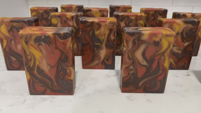 Rectangular bars of marbled artisan soap with swirls of orange, brown, cream, and yellow, standing upright on a white marble surface against a purple and magenta background.
